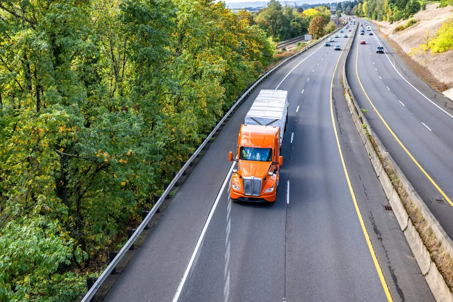 Orange semi-truck on a multi-lane highway surrounded by lush green trees on the left and dry grass on the right under a clear blue sky.